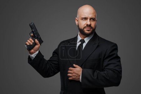 Foto de Hombre calvo y barbudo en un traje negro sostiene una pistola, exudando poder contra un fondo de estudio gris - Imagen libre de derechos