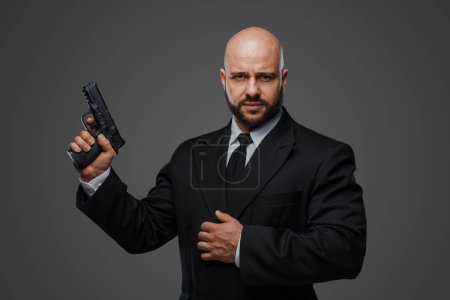 Téléchargez les photos : Homme chauve et barbu en costume noir tient un pistolet, exsudant puissance sur fond de studio gris - en image libre de droit