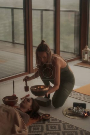 Téléchargez les photos : Une scène paisible de filles pratiquant des activités spirituelles et du yoga à l'aide d'un bol tibétain chantant, entouré d'une atmosphère apaisante dans un appartement avec une vue panoramique sur la ville - en image libre de droit