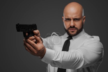 Téléchargez les photos : Homme chauve et barbu en chemise blanche et cravate vise un pistolet, canalisant une ambiance de détective ou de garde du corps dans un décor de studio gris - en image libre de droit