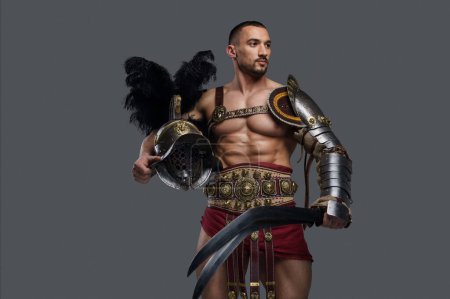 Téléchargez les photos : Gladiateur avec une barbe bien entretenue et une armure décorée légère pose avec un gladius et un casque à plumes sur un simple fond gris, rayonnant de force et de courage - en image libre de droit
