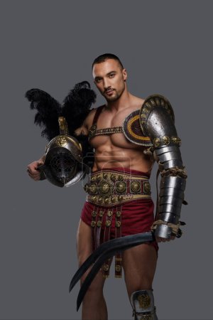 Téléchargez les photos : Gladiateur formidable avec une barbe élégante porte une armure légère au design complexe, tenant un gladius et un casque à plumes alors qu'il pose avec confiance sur un fond gris - en image libre de droit