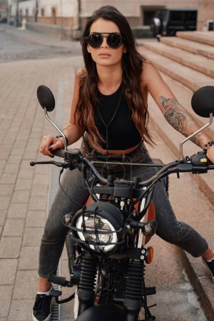Foto de Elegante morena en jeans y una camiseta sin mangas, con gafas de sol y un tatuaje en el brazo, posa en su motocicleta retro en una vieja calle empedrada de Europa - Imagen libre de derechos