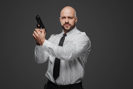 Téléchargez les photos : Homme chauve et barbu en chemise blanche et cravate tenant un pistolet, incarnant un détective, un garde du corps ou un professionnel de la sécurité sur fond de studio gris - en image libre de droit