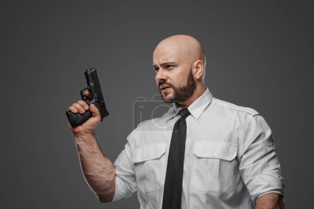 Foto de Hombre calvo y barbudo con camisa blanca y corbata sostiene una pistola, personificando a un detective, guardaespaldas o profesional de seguridad contra un fondo gris del estudio - Imagen libre de derechos