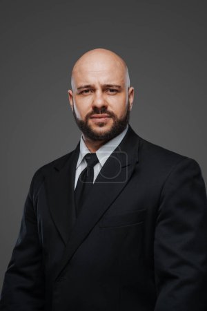 Photo for Authoritative bald gentleman with a stout build and beard, clad in formal black attire, set against a muted studio background - Royalty Free Image