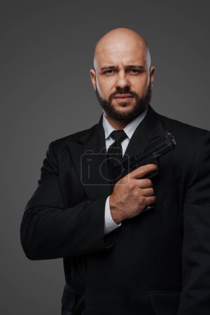 Photo for Bald and bearded man in a black suit holds a pistol, exuding power against a gray studio backdrop - Royalty Free Image