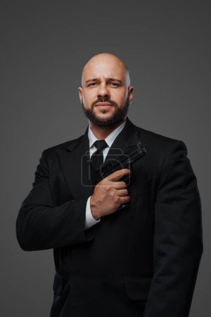Photo for Bald security man in a suit holds a gun, ready to protect, against a gray studio backdrop - Royalty Free Image