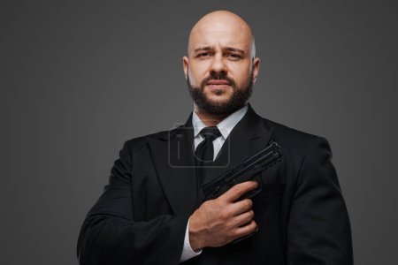 Téléchargez les photos : Un agent de sécurité chauve en costume tient une arme, prêt à protéger, dans un décor de studio gris - en image libre de droit