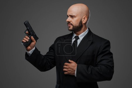 Foto de Calvo hombre de seguridad en un traje sostiene un arma, listo para proteger, contra un fondo de estudio gris - Imagen libre de derechos