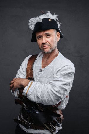 Photo for Mysterious pirate with a gray beard wearing a white shirt and hat, striking a pose with crossed arms against a textured gray wall - Royalty Free Image