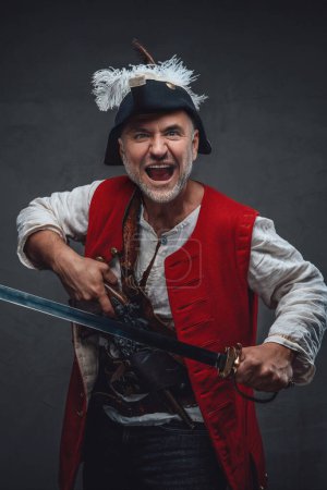 Foto de Un pirata amenazante con una barba gris vestida con un traje blanco y un chaleco rojo, sosteniendo un mosquete y una espada, preparándose para el combate contra un telón de fondo texturizado - Imagen libre de derechos