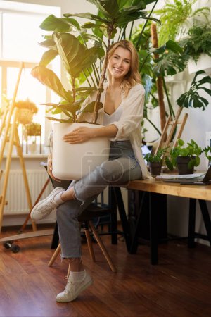 Téléchargez les photos : Un charmant fleuriste souriant tenant un grand pot avec une plante dans une pièce remplie de plantes et de lumière du soleil chaude provenant de la fenêtre - en image libre de droit