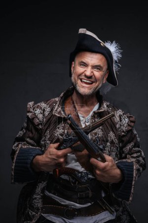 Foto de Un pirata de ojos salvajes con una sonrisa loca y barba gris vestido con un chaleco y sombrero sosteniendo dos mosquetes contra una pared oscura - Imagen libre de derechos