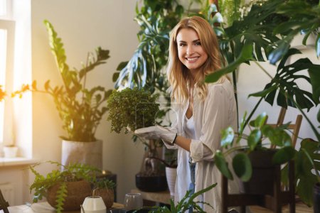 Photo for Happy florist holding a plant, surrounded by potted plants in a sunlit room - Royalty Free Image