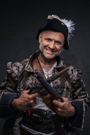 Foto de Un personaje pirata amenazante, envejecido y rugoso, luciendo una barba salvaje, chaleco y sombrero, blandiendo dos mosquetes frente a un fondo oscuro y texturizado - Imagen libre de derechos