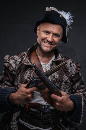 Foto de Un pirata de ojos salvajes con una sonrisa loca y barba gris vestido con un chaleco y sombrero sosteniendo dos mosquetes contra una pared oscura - Imagen libre de derechos