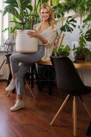 Téléchargez les photos : Fille souriante en chemise blanche et jeans présentant énorme plante en pot dans la pièce remplie de verdure - en image libre de droit