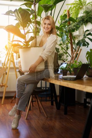 Photo for A cheerful florist in a white shirt and jeans holding a large potted plant in a sun-kissed room full of greenery - Royalty Free Image