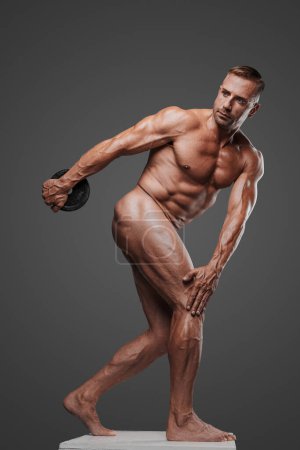 Foto de Un modelo masculino desnudo musculoso y atractivo sosteniendo una placa de peso, de pie sobre un pedestal en la pose de un atleta olímpico antiguo, rodeado de esculturas griegas antiguas, sobre un fondo gris - Imagen libre de derechos