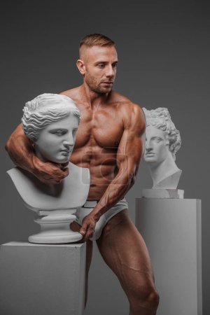Photo for A muscular, masculine man wearing only underwear poses alongside an ancient Greek bust on a grey background, exuding raw sensuality - Royalty Free Image