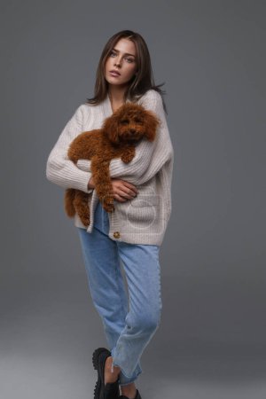 Full-length portrait of a captivating brunette in casual wear holding her brown toy poodle against a gray studio background