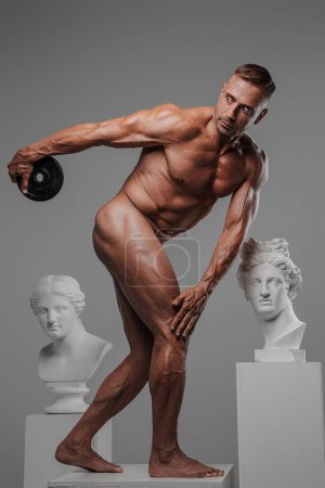 A muscular and attractive naked male model holding a weight plate, standing on a pedestal in the pose of an ancient Olympic athlete, surrounded by ancient Greek sculptures, against a gray background