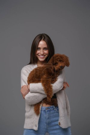Photo for Captivating young brunette with a wide smile cradling her cherished toy poodle, styled in a white top and denim shorts, before a gray studio background - Royalty Free Image