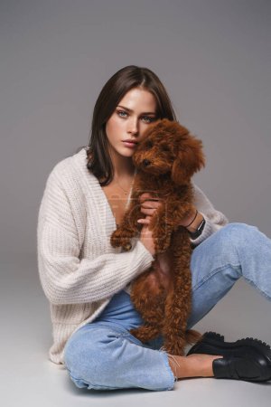 Photo for Elegant brunette in white top and denim shorts sits on the floor, adoringly holding her toy poodle, against a gray studio background - Royalty Free Image