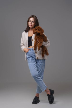 Photo for Full-length portrait of a captivating brunette in casual wear holding her brown toy poodle against a gray studio background - Royalty Free Image