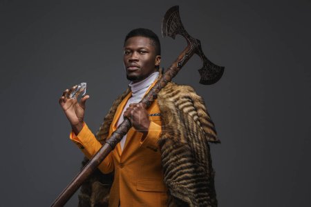 Photo for Dark-skinned man in a stylish yellow jacket, draped in the pelt of a wild animal holds massive diamond and axe against a neutral gray background - Royalty Free Image