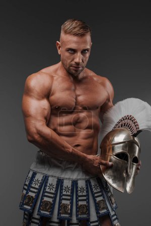 Téléchargez les photos : Un mannequin masculin musclé et séduisant vêtu de rideaux anciens et tenant un casque grec hoplite, posant sur un fond gris - en image libre de droit