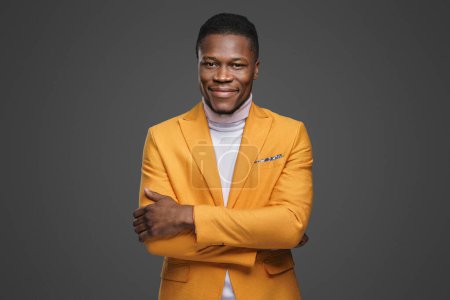 Photo for A handsome dark-skinned young man in an elegant yellow blazer and white turtleneck smiling and posing with crossed arms on a grey background - Royalty Free Image