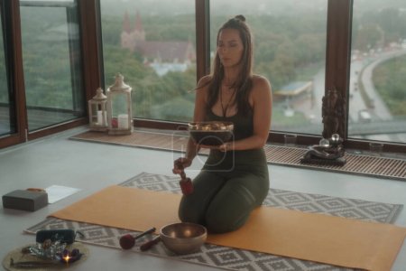 Photo for Woman in sportswear practicing spiritual activities in a peaceful environment, meditating with a singing Tibetan bowl in an apartment overlooking a panoramic city skyline - Royalty Free Image