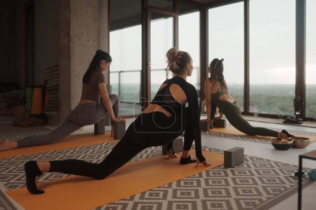 Téléchargez les photos : Groupe de femmes en vêtements de sport pratiquant le yoga et la méditation dans un appartement avec vue panoramique sur la ville - en image libre de droit