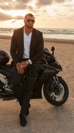 Téléchargez les photos : Homme élégamment habillé en costume noir s'appuie sur une moto de sport sur une plage désolée contre un coucher de soleil nuageux - en image libre de droit