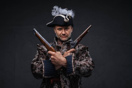 Photo for Seasoned pirate with a grey beard dressed in a brown vest posing with two single-handed muskets against a textured grey wall - Royalty Free Image