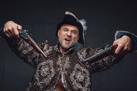 Photo for A wild-eyed pirate with a crazy smile and gray beard dressed in a vest and hat holding two muskets against a dark wall - Royalty Free Image