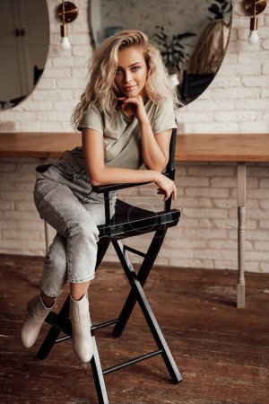 Téléchargez les photos : Élégante blonde avec une coiffure volumineuse posant sur une chaise dans une salle de maquillage - en image libre de droit
