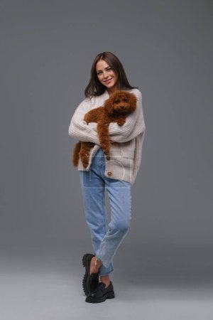 Photo for Radiant young brunette in casual wear beaming with happiness while holding her cherished toy poodle, set against a neutral gray studio background - Royalty Free Image