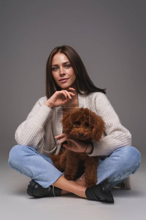 Photo for Attractive young brunette in white top and denim shorts, seated on the floor, cradling her brown toy poodle, against a gray studio backdrop - Royalty Free Image
