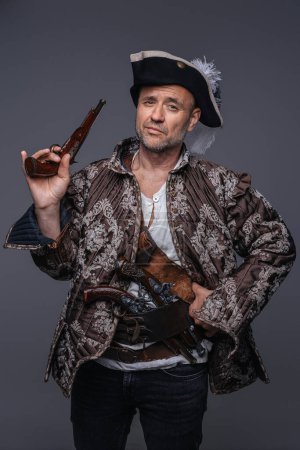 Photo for Pirate showcasing his seasoned look, wearing a brown vest and sporting two single-handed muskets against a textured grey wall - Royalty Free Image