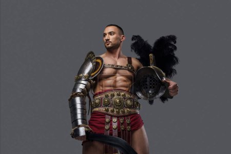 Foto de Gladiador muscular en elegante armadura ligera golpea una pose mientras sostiene un gladius y un casco, sobre un fondo gris - Imagen libre de derechos