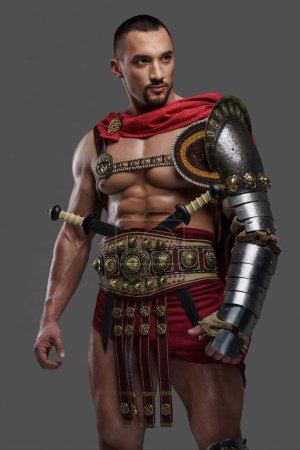 Foto de Potente gladiador con una elegante barba lleva una armadura ligera e intrincada y posa agarrando un casco emplumado sobre un fondo gris - Imagen libre de derechos