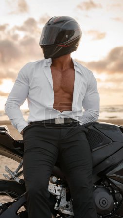 Téléchargez les photos : Motocycliste avec casque et chemise blanche ouverte, révélant un torse tonique, par son vélo noir, contre un coucher de soleil morose sur la plage - en image libre de droit