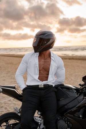 Photo for Motorcyclist with helmet and open white shirt, revealing a toned torso, by his black bike, against a moody beach sunset - Royalty Free Image