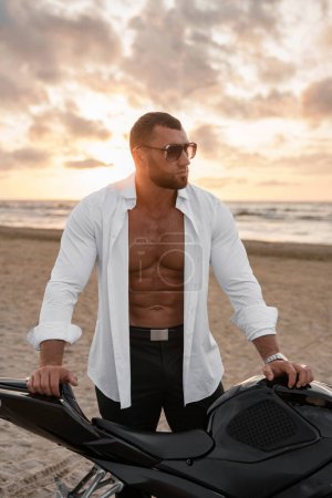 Striking man in sunglasses and open white shirt, showcasing a toned torso, stands by his black motorcycle, desolate beach and cloudy sunset behind