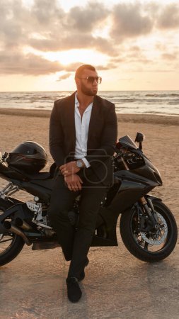 Téléchargez les photos : Homme élégamment habillé en costume noir s'appuie sur une moto de sport sur une plage désolée contre un coucher de soleil nuageux - en image libre de droit