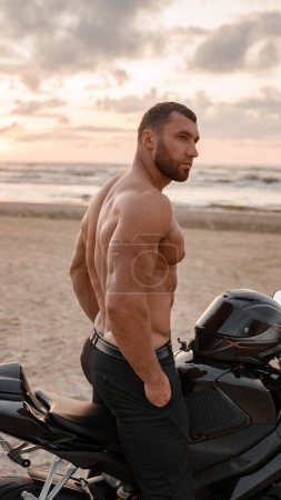 Téléchargez les photos : L'homme musclé à son apogée est assis en toute confiance sur son vélo de sport noir sur un rivage vacant, avec l'allure d'un coucher de soleil nuageux derrière - en image libre de droit