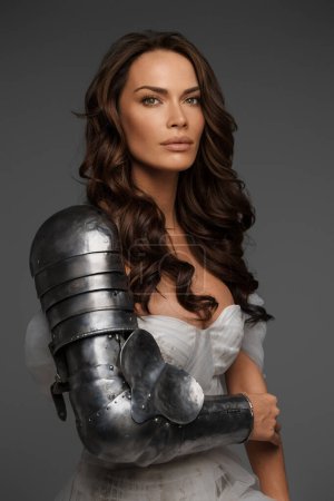 Photo for Beautiful middle-aged model in white dress and knights armor poses gracefully on gray - Royalty Free Image